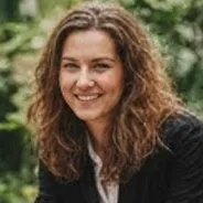 Smiling woman with curly brown hair in business clothes, warm outdoor light and lush green foliage background