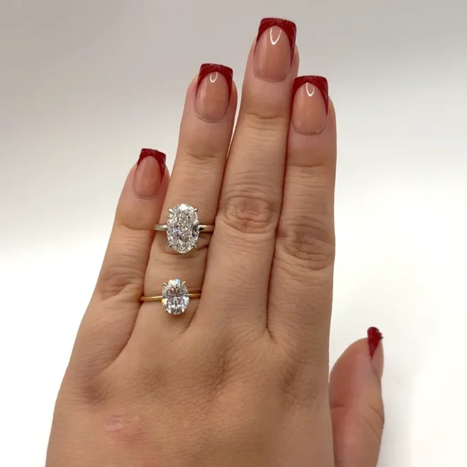 Glossy red French-tip manicure on an elegant hand showcasing two oval-cut diamond rings set in gold bands against a clean white background.