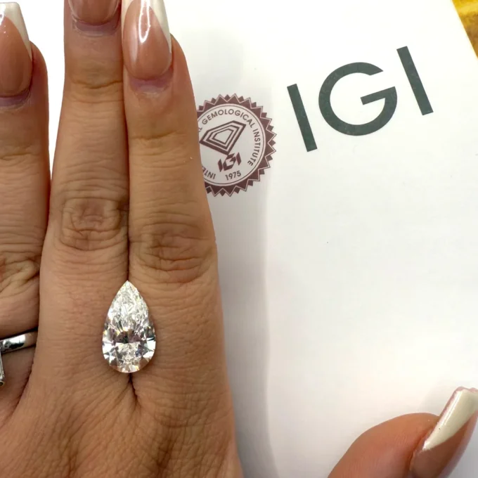 Pear-shaped diamond engagement ring on a manicured hand beside an IGI certificate — a brilliant teardrop solitaire showcased with polished nails.
